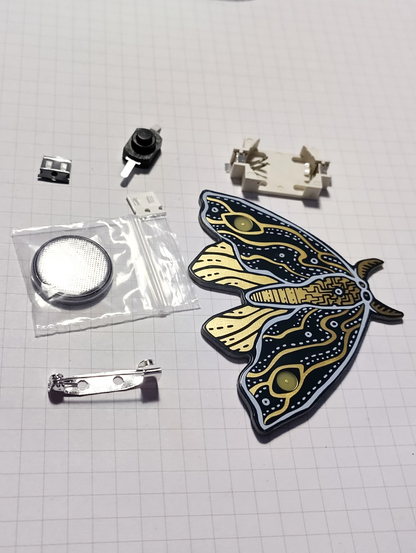 A flat-lay shot of a partially assembled moth-shaped PCB (Printed Circuit Board) kit on a light grid background. The PCB is dark with gold and white patterns resembling abstract designs, showing visible electronic traces. To the left of the PCB are several small components: a surface-mount button, a through-hole button, a clear plastic bag containing a round, silver battery, a small metal battery holder, and a silver safety pin. In the upper right corner, there is a white plastic component, possibly a battery holder or connector. The components are neatly arranged, indicating a DIY or assembly kit.