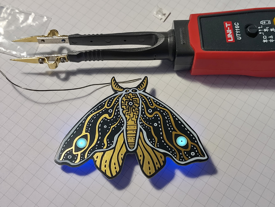 The photo shows an assembled moth-shaped PCB lying on a light grid background. The moth has a black body with gold and white patterns, and its eyes are illuminated with blue LEDs. In the upper part of the photo, there is a red UNI-T UT116C multimeter with black probes ending in gold tips, which are pointed downwards towards the moth. A thin black wire comes out of the lower part of the multimeter probes and runs towards the moth. In the upper left corner, there is a small, transparent bag with small components.