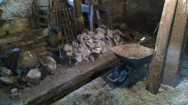 This image shows a rustic barn or stable with dirt-covered floors and aged, concrete walls. An emty wheelbarrow near a wooden support pillar is located in the middle of the image. Stones are scattered around, indicating ongoing work. Black iron bar and pick leaning against the back wall.