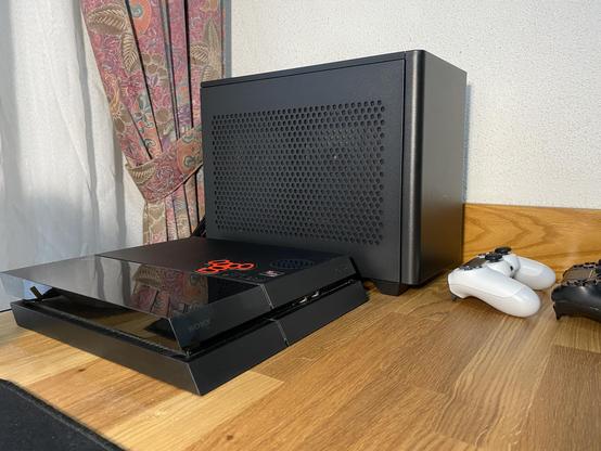 Imagen en la que se ve un ordenador de sobremesa en formato mini-ITX junto a una PlayStation 4 de color negro y sus dos mandos DUALSHOCK 4. Uno de color blanco y otro de color negro.
