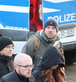 Foto von der Neonazi-Demo am 15.02.25 in Dresden. Vor einem Polizeiwagen ist Jan Tiemann in dunkelblauer Mütze und hellgrauer Jacke zu sehen. Er trägt einen olivgrünen Rucksack.   Bild von Tim Mönch