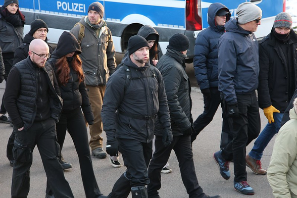 Foto von der Neonazi-Demo am 15.02.25 in Dresden. Im hinteren linken Bildteil ist vor einem Polizeiwagen Jan Tiemann in dunkelblauer Mütze, hellgrauer Jacke und brauner Hose zu sehen (links daneben Jan Steffen Holthusen). Er trägt einen olivgrünen Rucksack.   Bild von Tim Mönch