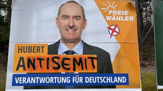 Das Bild zeigt ein Wahlplakat der "Freien Wähler". Das Gesicht des Kandidaten (Hubert Aiwanger) wurde mit einem Hitler-Bart versehen. Über dem Slogan "Verantwortung für Deutschland" und nach dem Vornamen "Hubert" wurde mit Farbe das Wort "Antisemit" gesprüht. Das Logo der "Freien Wähler" mit einer stilisierten Sonne und einem durchgestrichenen Wahlkreuz ist ebenfalls zu sehen. Das Plakat hängt im Freien, wahrscheinlich in einem ländlichen Gebiet.

Bereitgestellt von @altbot, generiert mit Gemini