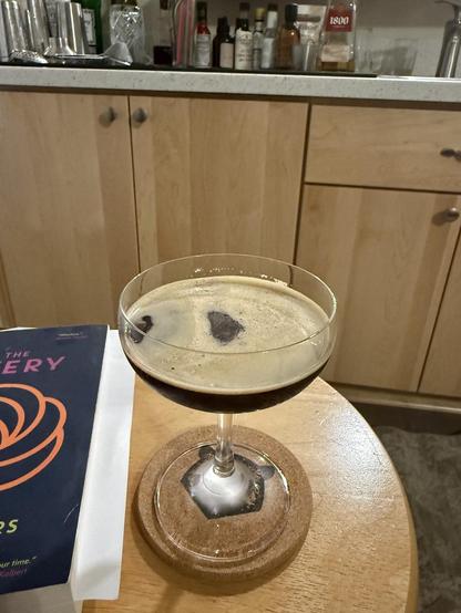 An espresso martini in a coop glass sits a top of cork coaster that’s on a round wooden table. In the background is a home bar and beside the drink is a book, ready to be enjoyed.