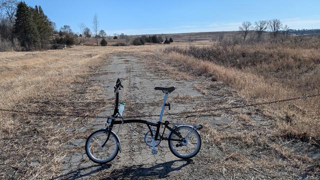 Black and silver Brompton C-line bicycle leaning on a chain gate line at the end of a beat up asphalt road heading off to vacant land with brown grass and scrubby trees. The sky is blue.
