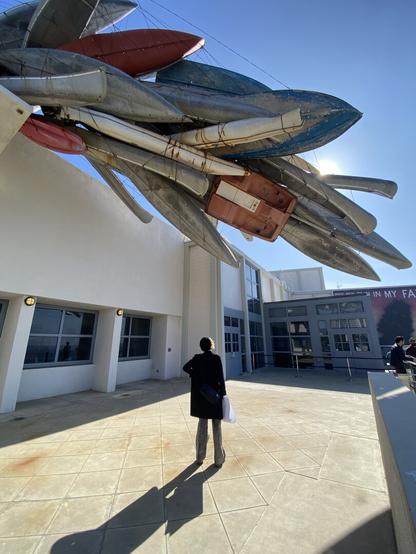 A person standing under a statue made of apparently a dozen boats, mostly metal, suspended above. The sun is partially visible behind the sculpture.