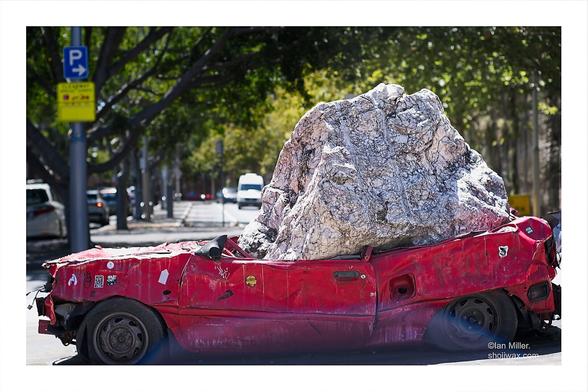 Art installation of a crushed red car with a huge quartz bolder on top of it.