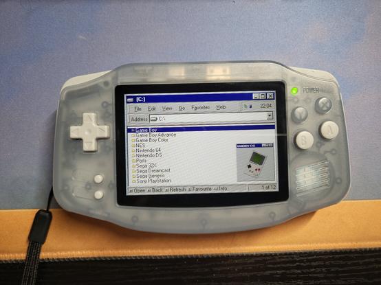A Retro Handheld shaped like the original Game Boy Advance, displaying a screen that resembles the old File Explorer from Windows 98. It is displaying a list of systems, and the highlighted system (the original Game Boy) has a picture that looks like a pop up window.