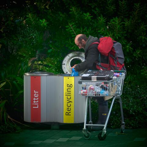A person stands next to labelled bins named 'Litter' and 'Recycling'. They are placing an item in the recycling bin and have a shopping cart filled with various objects. The scene is set outdoors with green foliage in the background.