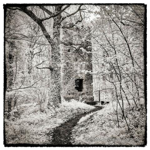 A toned black and white infrared photo of a ruined brick tower standing in woodland, with a path leading towards it.