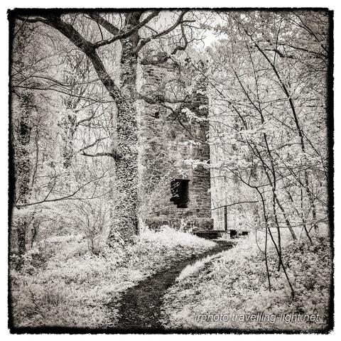A toned black and white infrared photo of a ruined brick tower standing in woodland, with a path leading towards it.