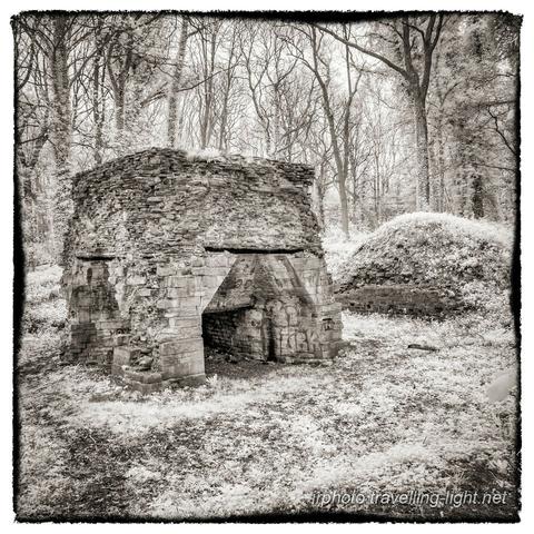 A toned black and white infrared photo of a brick and stone furnace standing in woodland.