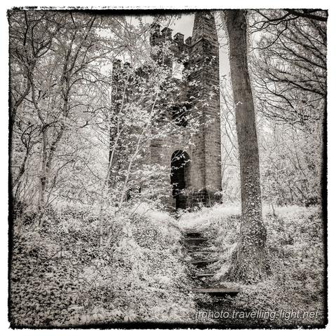 A toned black and white infrared photo of a ruined brick tower standing in woodland, with a path leading up steps towards it.