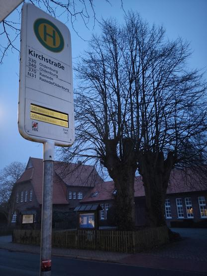 Foto einer Anzeige an einer Bushaltestelle. Im Hintergrund ein dörfliches Gemeindezentrum in der Morgendämmerung.