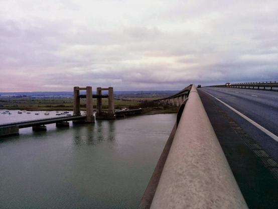 A view from the Sheppey Crossing bridge looking across a body of water, with the concrete pillars of a parallel bridge visible to the left. The image is taken from the perspective of standing on the bridge's edge, with the concrete barrier running along the right side of the frame. In the distance, a vehicle's lights can be seen on the curved roadway. The scene is captured under an overcast sky with moody gray clouds stretching across the horizon. Small boats can be spotted in the water below, and the landscape extends into flat marshland in the background.