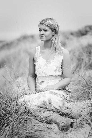 Junge Frau sitzt in einem Kleid in den Dünen und schaut in die Ferne