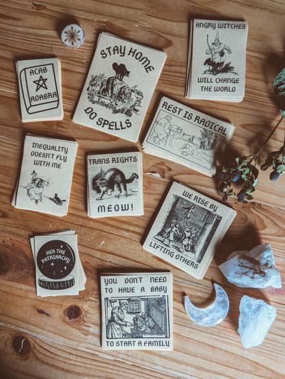 Mehrere Stapel Sticker liegen auf einem Holztisch. Sie zeigen unterschiedliche Motive wie Hexen, Katzen usw. Dazu Sprüche wie:

- Rest is radical

- You don’t need to have a Baby to start a family

- Angry Witches will Change the World

- Trans Rights Meow

- We rise by Lifting Others

- acab adabra

- stay Home do spells

- Inequality doesn’t fly with me

- Hex the patriarchy