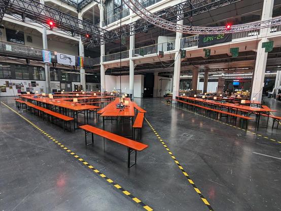 Das Bild zeigt eine große Veranstaltungshalle mit hohen Decken und mehreren Stockwerken. In der Mitte sind lange, orangefarbene Biertischgarnituren (Tische mit dazugehörigen Bänken) aufgestellt, einige mit kleinen Lampen und Kabeln darauf. Der Boden ist mit schwarz-gelb gestreiftem Klebeband markiert, vermutlich zur Abgrenzung bestimmter Bereiche.

Im Hintergrund sind weitere Arbeitsplätze mit Stühlen, Laptops und technischem Equipment zu sehen. Eine digitale Anzeige an der oberen rechten Seite des Bildes zeigt den Text „GPN-22“, was auf eine Veranstaltung oder Konferenz hindeuten könnte. Zudem hängen an den Geländern Banner, darunter eine Regenbogenflagge und ein Banner mit einer stilisierten Zeichnung einer Person.

Die Szenerie deutet auf eine Hackerkonferenz oder eine technische Veranstaltung hin, möglicherweise ein Hackathon oder eine LAN-Party. Die Atmosphäre ist gut organisiert, aber noch relativ leer, was darauf hindeuten könnte, dass die Veranstaltung noch nicht begonnen hat oder gerade erst startet.