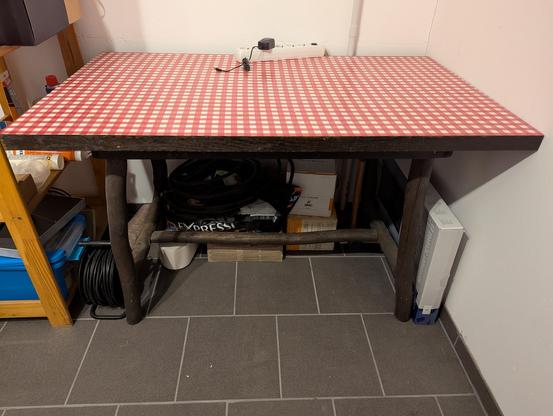 Foto von einem massiven Tisch mit rot-weiß kariertem Furnier. Der Tisch steht in einem Keller und ist braun.