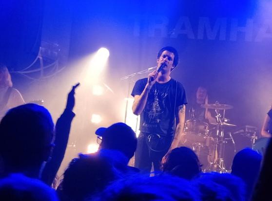 Le chanteur de Tramhaus, Lukas Jansen, micro en main, sur le devant de la scène du Petit Bain, face à son public que l'on voit au premier plan. Le batteur est en arrière plan. Lumière bleutée avec éclairage puissant de spot.