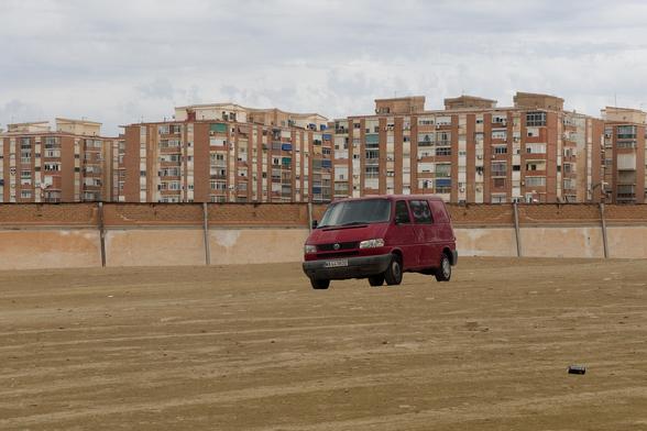 A solitary red van sits adrift on a wide, dusty expanse, as if it’s strayed from a distant road trip. Looming in the background, rows of worn apartment blocks stand like silent spectators, their uniform balconies forming a stark contrast to the barren ground below. The overcast sky adds a muted hush to the scene, highlighting the van’s unexpected cameo in this urban desert.