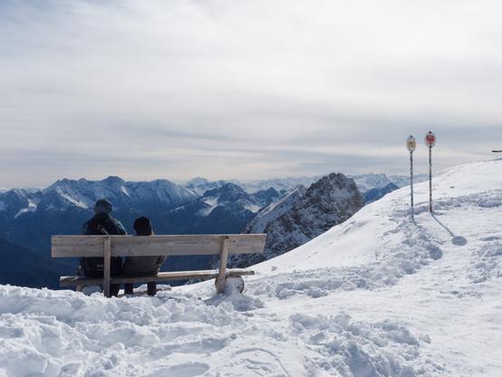 A couple sits on a wooden bench atop a snow-covered mountain, gazing at the breathtaking alpine landscape. In the distance, rugged peaks stretch into the horizon beneath a pale winter sky. Nearby, two signposts mark the border between Germany (Bavaria) and Austria (Tyrol), their poles covered in stickers left by previous visitors. The untouched snow and crisp mountain air create a serene and timeless moment of quiet reflection.