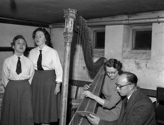 A  black and white photograph from 1958 of a woman playing a harp while looking at a sheet of music held by a man, with two women singing the Cerdd Dant at a festival