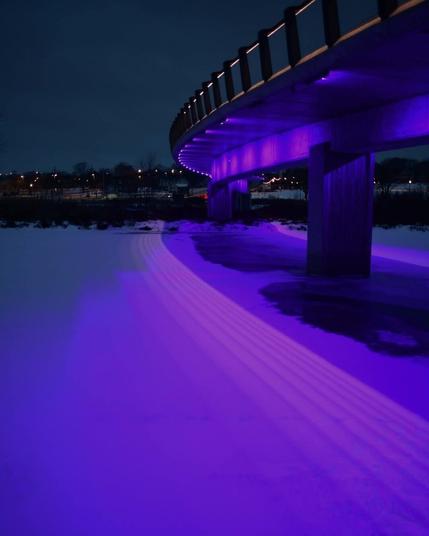A very purple-hued image, tinted from the colors of an early rising sun. 

A frozen river is covered in snow, with the thin outline of city buildings visible across the far distance. A few open spots in the ice can be seen underneath a curved bridge. The bridge is held up by thick cement supports and very gently highlighted with regular small lights. It curves and swoops gracefully from the right side, and disappears into the city behind it. The bridge looks very large compared to the city, giving the illusion that it is incredibly long (it isn't). 

Photo credit: Bill Peterson