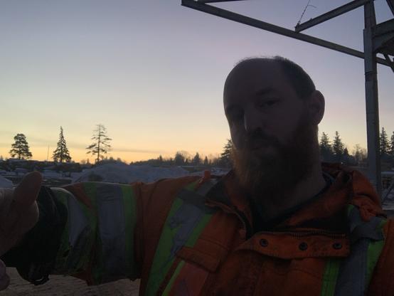 A large cis white male with a red beard offers out a hug. He’s wearing a reflective jacket but is in a shadow