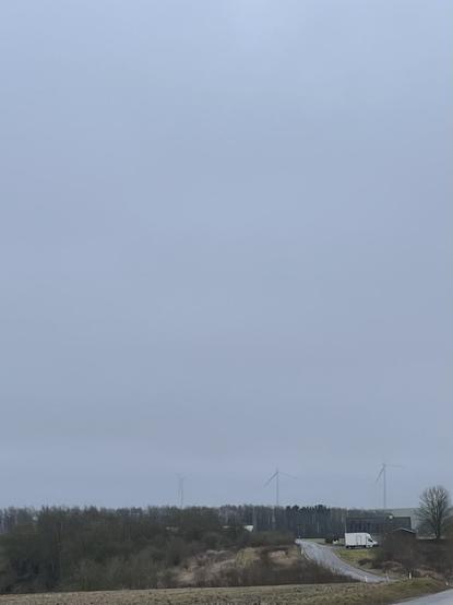 Foto ud over et landskab. I forgrunden en grøn mark og en vej med en lastbil parkeret ved siden af, vejen fører op til et hegn af træer og bag dem står tre vindmøller med hver af de øverste vinger væk i dis og en grå himmel, som fylder de øverste tre fjerdedele af billedet.