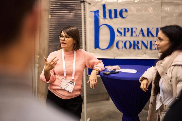 Julijana Gjorgjieva stands at a high desk, speaking to people gathered around her at the blue speakers' corner.