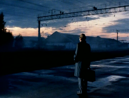 Un hombre con un abrigo oscuro y un sombrero fedora de pie de espaldas en una plataforma de tren vacía, mirando hacia un cielo crepuscular.  Al fondo, se ve una línea de edificios industriales bajo un cielo anaranjado y gris, con una estructura de catenaria de ferrocarril elevándose sobre la escena. El hombre lleva un maletín. La atmósfera es sombría y evocadora.