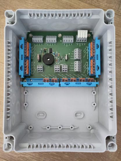 Photo du prototype en gros plan : une boîte en plastique gris, ouverte sur le dessus. Au fond de la boîte est fixé un circuit imprimé vert sur lequel sont soudés des composants (résistances, etc.) On voit aussi des borniers gris soudés, et des borniers transparents avec levier orange qui sont généralement utilisés dans les installations électriques domestiques, mais ici ils sont fixés au circuit par l'intermédiaire de supports bleus. Des indications sont imprimées sur le circuit pour savoir où les fils doivent être branchés (mais sur cette photo il n'y a aucun fil raccordé au boîtier)