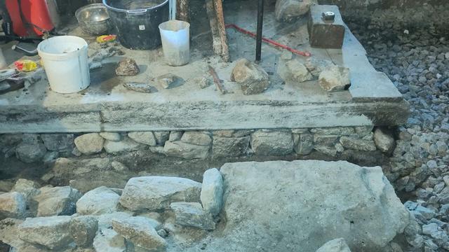 This image shows the construction or renovation of a manure channel in a stable. The lower section features a partially exposed stone foundation, while the upper section has a wooden platform. Various tools and materials, including buckets, stones, and a red-handled tool, are present, indicating active masonry work. The channel appears to be undergoing repair to improve the durability.