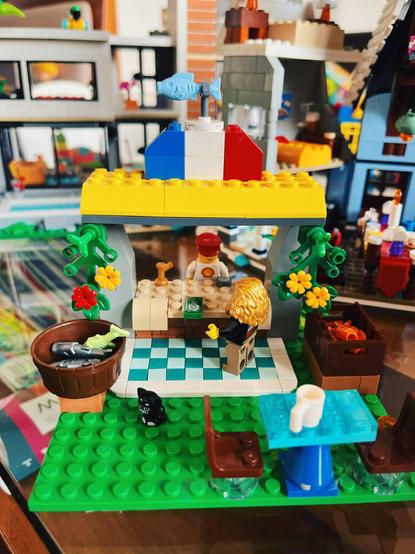 A photo of an original LEGO creation with a stand and a man with a man with a red cap behind it. There is a French flag on top and a fish sign. Below you see a long-haired customer standing on checkered square tiles. On the left there is a bucket with grey and green fish and on the right a brown case full of orange fish. In the foreground there is a small table with a white mug on it and two brown chairs on opposite ends.