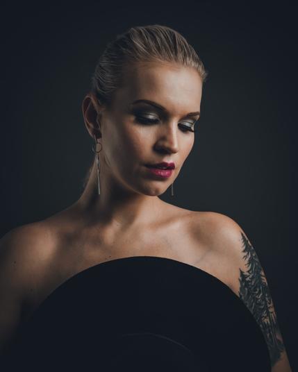 A close-up portrait of a woman looking upward towards the light. Her hand is resting behind her head, creating a strong yet elegant pose. The lighting emphasizes her glowing skin and sharp facial features against a dark studio background. Valokuvaaja Kuvaajankulma Jarno.S Lappeenranta Suomi