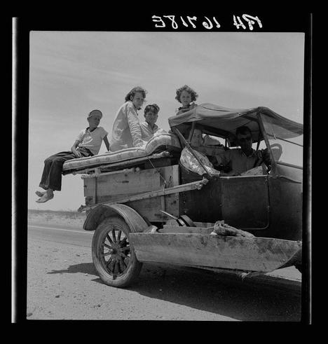 A black and white photograph depicting a group of five individuals in the backseat of an old, makeshift vehicle. The scene captures them traveling across a desert landscape under bright sunlight. One person is seated on a mattress placed atop the wooden structure of their car, while another leans against it with legs crossed. A child sits between two adults who are sitting close to each other; one appears relaxed with arms resting behind him and his feet elevated in the air. The driver's side shows an adult man wearing sunglasses driving attentively. There is also a sleeping dog on the wooden platform beside them, contributing to this candid snapshot of their journey.