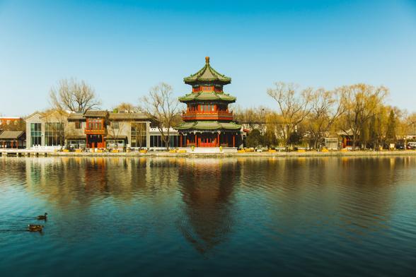 A red pagoda with green roof tiles sits in the center of the photo, a blue sky above, fading into a very light blue, to almost white behind the pagoda. Buildings line the street to the left and right of the pagoda and trees also grow in front of those buildings. A big lake is in the foreground with 2 ducks swimming past