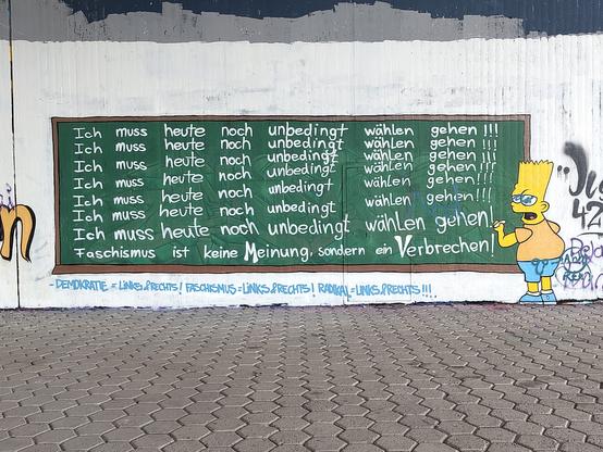 Graffiti:
Bart Simpson schreibt mit weißer Kreide auf eine grüne Tafel sieben Mal den Satz "Ich muss heute noch unbedingt wählen gehen !!!" untereinander. Darunter steht der Satz "Fschismus ist keine Meinung, sondern ein Verbrechen!"