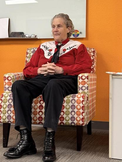 Temple Grandin, sitting in a chair during introductions. She’s wearing a red western-themed shirt, black pants, and boots.