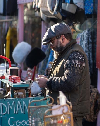 A masc person with an impressive beard, in a grey cap, green gillet and patterned knitted jumper. They are working at their stall.
