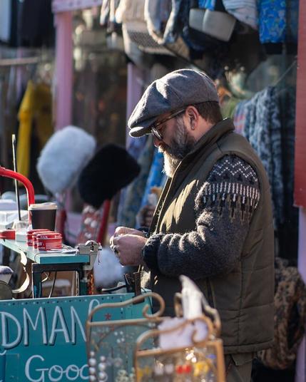 A masc person with an impressive beard, in a grey cap, green gillet and patterned knitted jumper. They are working at their stall.