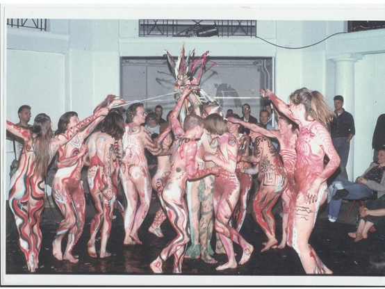 A neonaturist body paint performance by a cluster of young women dancing around a Maypole. They are painted with a variety of red, white and black squiggles and stripes