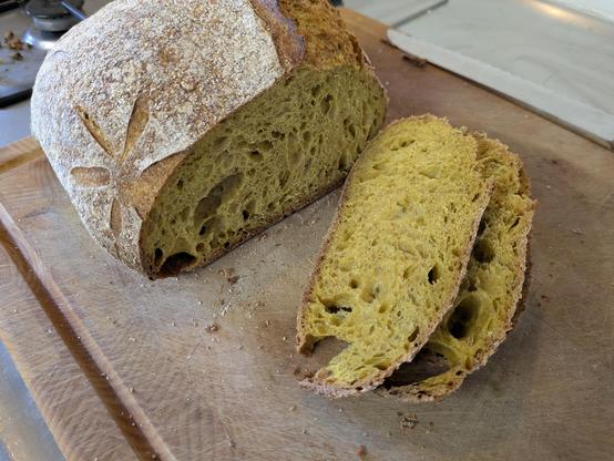 A partially sliced loaf of sourdough bread on a wooden chopping board. The crumb has an unusual yellow colour.