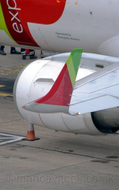 Close-up of the left upturned wingtip of a white, twin engined jet airliner parked facing to the left.
The wingtip has red, light green, and dark green stripes, and is extremely faded towards the rear edge.
The engine pods are white, and slung low under the wings.
There is a light green and red logo on the plane's forward fuselage, with the white letters "exp" overlaid vertically.
There are black "Operated by Portugalia Airlines" titles just forward of the wing root.
Grey concrete apron fills the rest of the bottom of the frame.