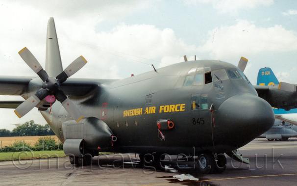 Front side view of a green, high-winged, 4 propellor-engined military transport aircraft parked facing to the right.
There are bright yellow "Swedish Air Force" titles on the forward fuselage, just above the number "845" in black below the cockpit windows.
There is an open door hanging down from the forward fuselage on the opposite side, visible under the nose.
A puddle runs along the concrete taxiway under the plane.
The blue and yellow tail of a similar plane can be seen in the background on the right of the frame.
Grey sky fills the rest of the frame.