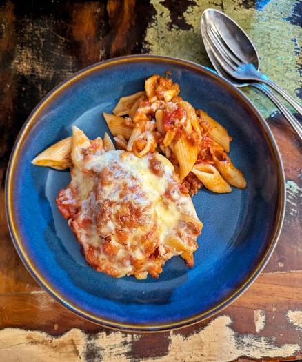 Dunkler Holztisch mit hellen Farbflecken. Darauf ein tiefblauer Steingutteller mit braunem Rand. Daneben eine in einen Löffel gelegte silberne Gabel. Im Teller Nudelauflauf mit Bolognesesauce und Käse.