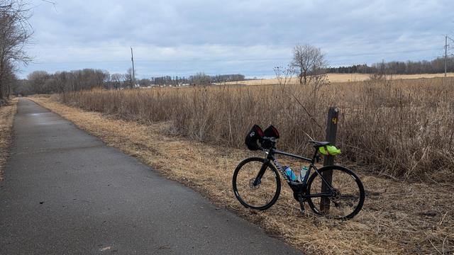 Blue and black Roubaix bicycle leaning on the wooden, brown painted, milepost "4" on the Douglas State Trail just South of Douglas looking North. The wet asphalt trail is on the left going straight, there are tall, dead weeds on the right and a lighter brown far field in the distance on the right. There are houses and trees center distance. The sky is mostly cloudy.