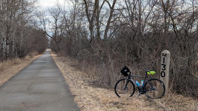Blue and black Roubaix bicycle leaning on the concrete obelisk Chicago Great Western Railway milepost "M130" (or 130 miles on that line to Mankato). The partially wet asphalt trail is on the left going straight, looking Southeast. The trail is surrounded by bare trees. The sky is cloudy.