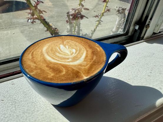Small blue mug filled with a frothy cappuccino sitting on a window sill with a rose bush in the background.